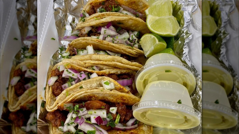 A row of tacos with sauces and limes in tin foil and styrofoam container