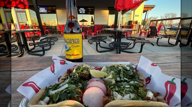 Multiple tacos on metal tray in outdoor seating area with pacifico beer bottle on wooden table