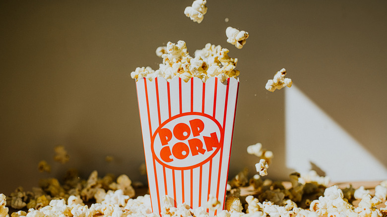 Small popcorn bucket filled with popcorn and additional pieces falling into the bucket, while popcorn surrounds on a table surface.