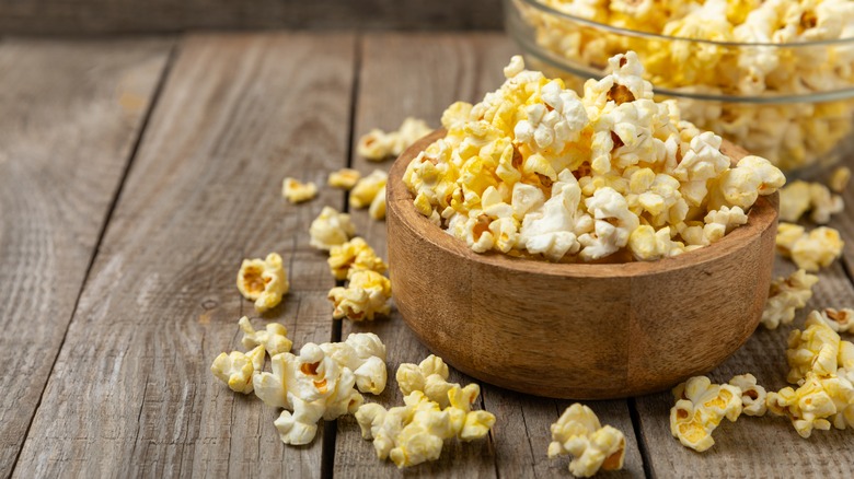 Two bowls of popcorn with popped kernels spread around on the wooden table underneath.