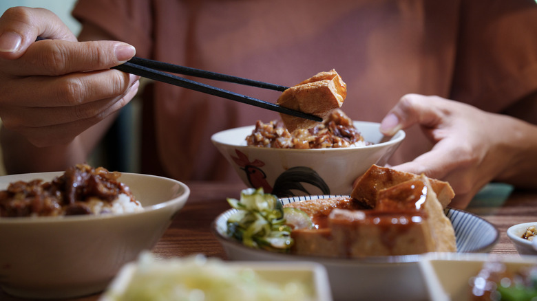 Hands holding a bowl of rice and chopsticks with various side dishes scattered around.