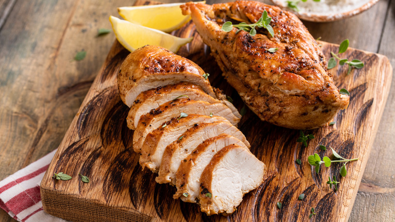 Sliced and whole cooked chicken breasts on wooden board next to lemon wedges