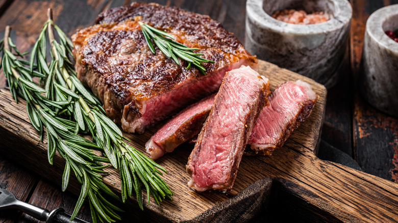 Grilled ribeye with a few slices on wooden board next to sprigs of rosemary