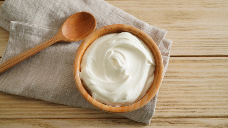 Sour cream in a wooden bowl next to a wooden spoon on top of a cloth napkin and wooden surface