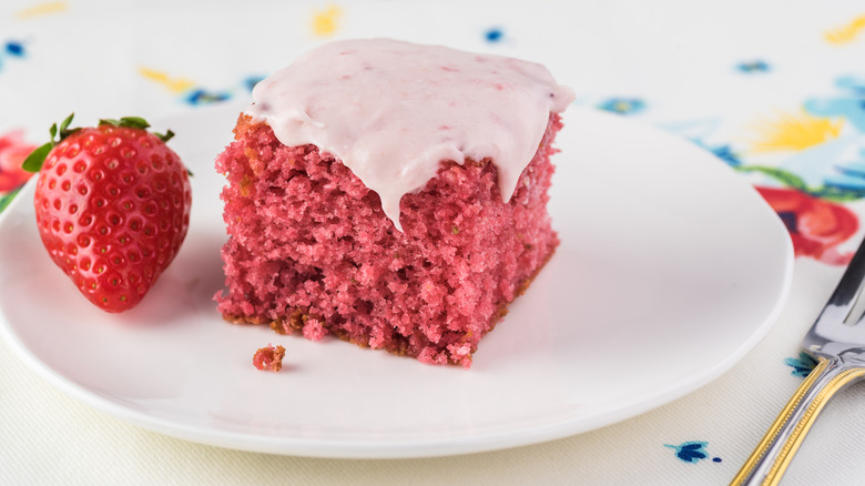 Square piece of pink strawberry cake with light pink frosting on a round white plate with a strawberry