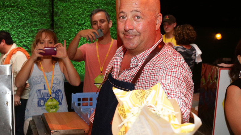Andrew Zimmern offering wrapped up food to the camera with people taking photos behind him
