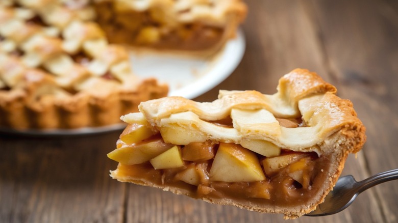 A slice of apple pie on a cake cutting knife with a partial whole pie in the background