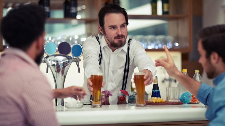 A bartender serving two beers to two customers as one of them points his finger up