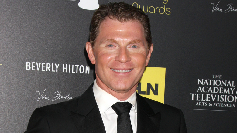 Close-up of Bobby Flay wearing a black suit and tie and white shirt against a dark promotional background