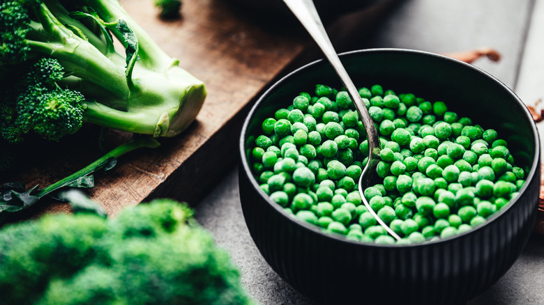 Frozen peas in a black bowl with a spoon and some fresh broccoli on a wooden board