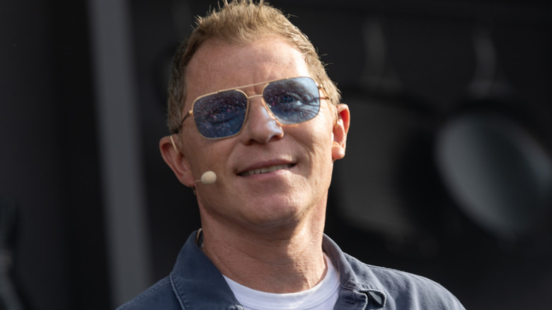 Bobby Flay wears blue sunglasses and a mic on stage at an event