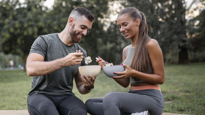 Athletic man and woman sitting outside eating from bowls while smiling