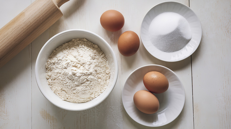 A wooden table that has eggs, sugar, and flour on it with a rolling pin nearby