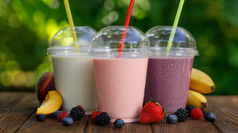 White, pink, and purple smoothies in plastic cups with tops and straws, surrounded by fresh fruit