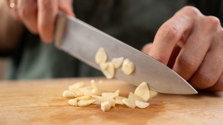 Male chef slicing larger slices of garlic cloves on a wooden cutting board with a big knife, close-up.