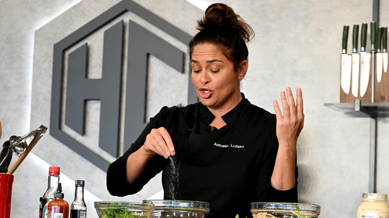 Chef Antonia Lofaso mid-demonstration, sprinkling seasoning into a glass bowl while speaking. Bottles of soy sauce and sriracha sit on the table, with a knife set visible in the background.