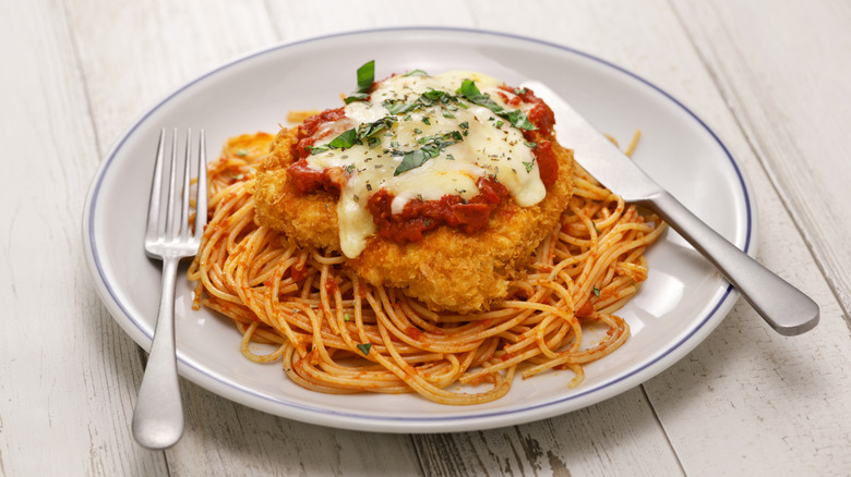 Breaded chicken parmesan on top of spaghetti noodles on a white and blue plate with a fork and knife also on it