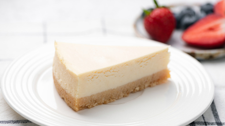 A slice of plain cheesecake with a graham cracker crust on a solid white glass dish and strawberries and blueberries in the background