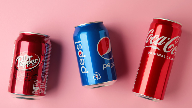Cans of Dr Pepper, Pepsi, and Coca-Cola on a pink background.
