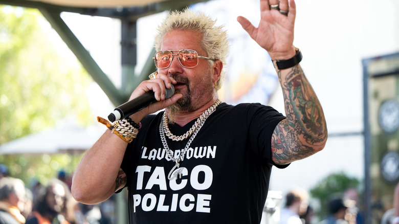 Guy Fieri in sunglasses waving and speaking into microphone
