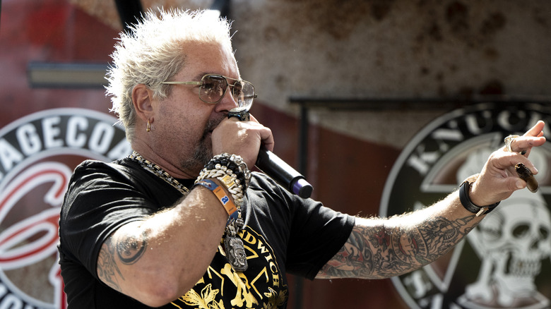 Guy Fieri speaking into microphone on stage at an event.