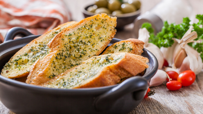 Sliced garlic bread with green herbs in black bowl with garlic, tomatoes, parsley, and olives in the background
