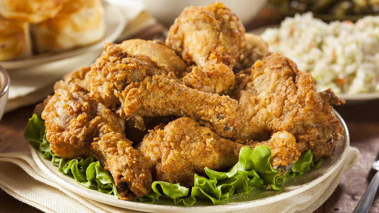 A plate with pieces of seasoned fried chicken on a bed of lettuce with sides in the background