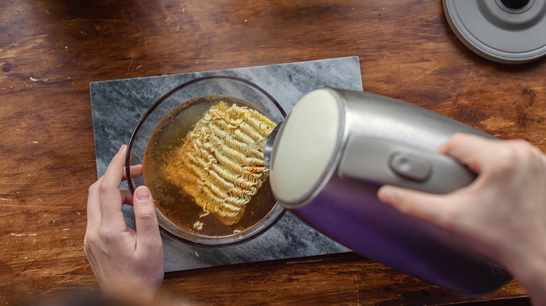 Hands pouring hot water from an electric kettle while preparing instant noodle soup.