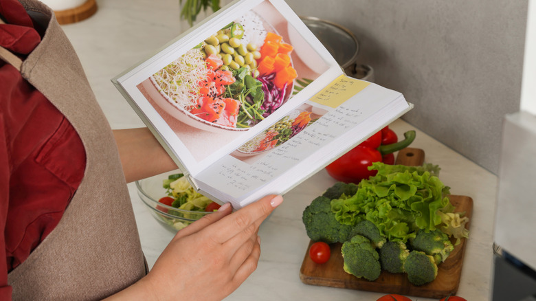 person wearing an apron gazing down at book with recipe in it and prepped fresh veggies in front of her