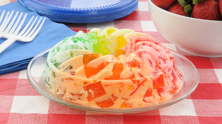 A multi-colored Jell-O dessert on a checked tablecloth near napkins, plates, forks, and a bowl of strawberries.