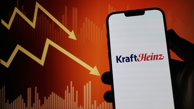 A hand holds a smartphone showing the Kraft Heinz logo against an image of stock market indicators in decline