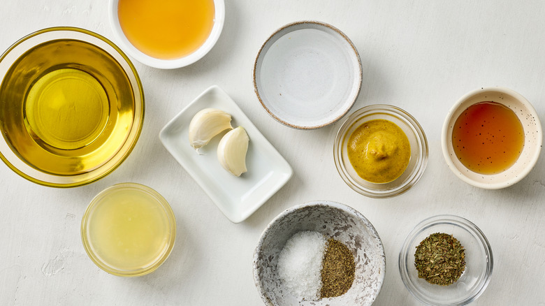 A view of vinaigrette ingredients on a white surface, including bowls of olive oil, vinegar, lemon juice, mustard, herbs, salt and pepper, and garlic cloves. 