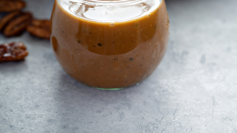 A glass jar of vinaigrette with a whisk inside, set on a surface with a green salad in the background.
