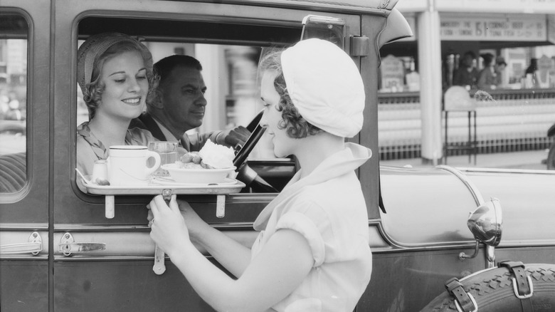 A black-and-white image of a carhop delivering food to a couple in a car.