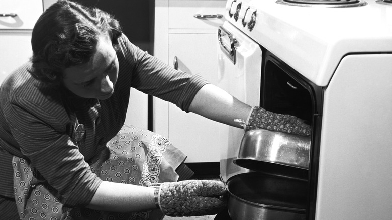 A vintage photograph of a cook putting a casserole dish in an oven.