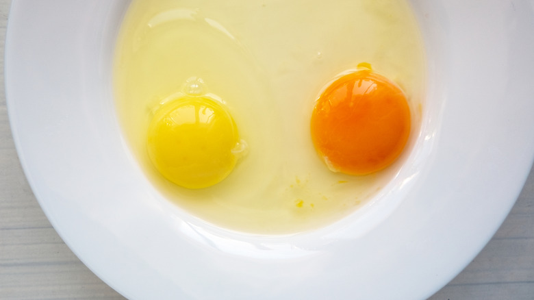 Two cracked raw eggs on white plate, one with a yellow yolk and one with an orange yolk