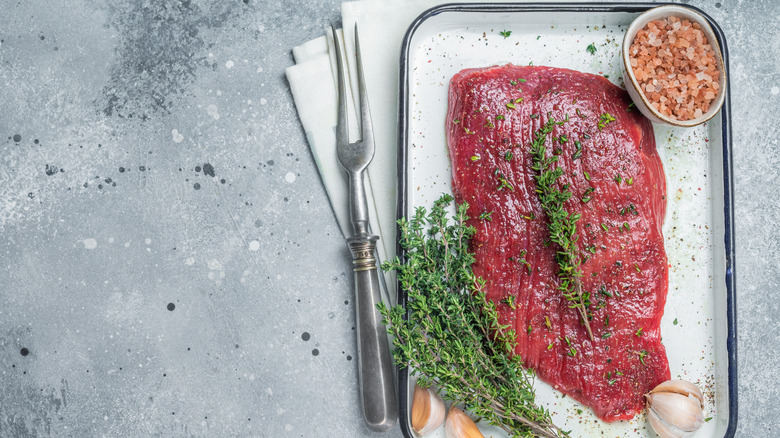 Uncooked steak on pan with fresh herbs, garlic, rock salt