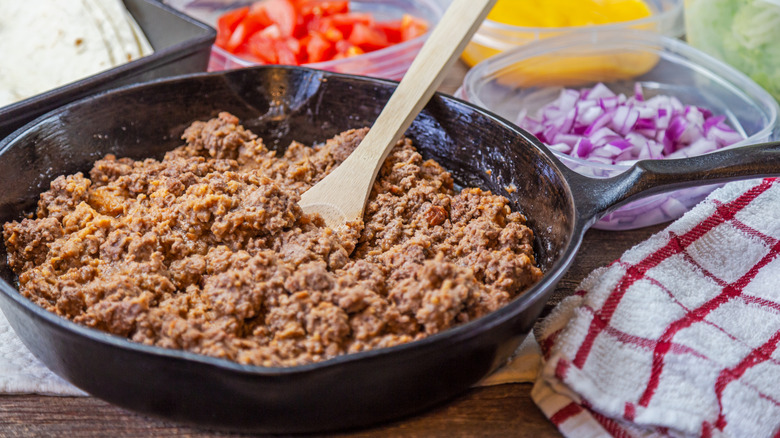 Ground beef in a skillet near items for tacos in small plastic bowls outside on a picnic table.
