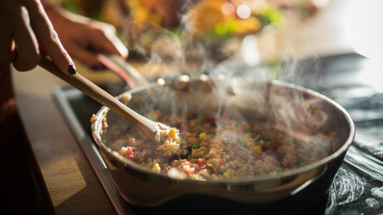 hand holding wooden utensil stirs steaming pan of rice and vegetables
