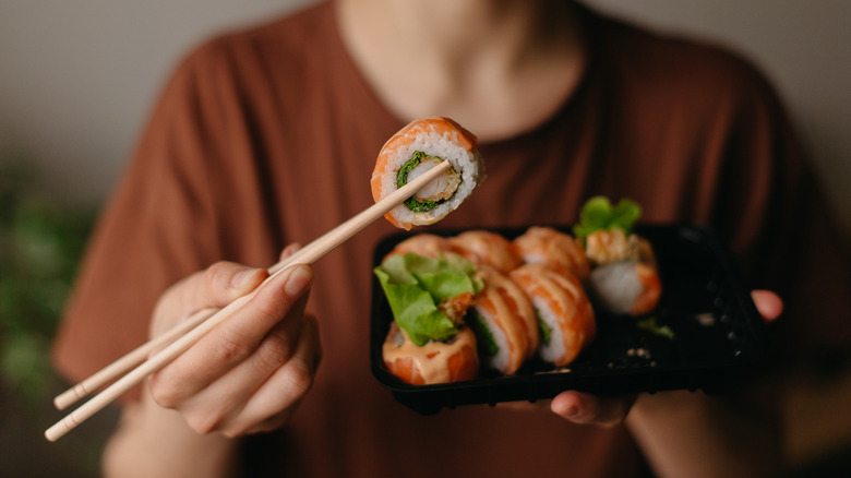 Someone holding up salmon sushi roll piece with a pair of wooden chopsticks