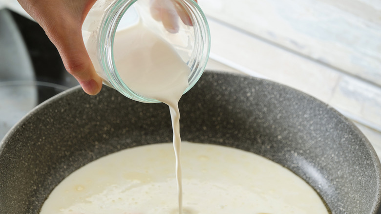 Milk being poured into a cream-based sauce in a pan on the stovetop