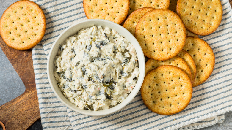 A white ramekin containing spinach dip on a cloth napkin and cutting board next to crackers