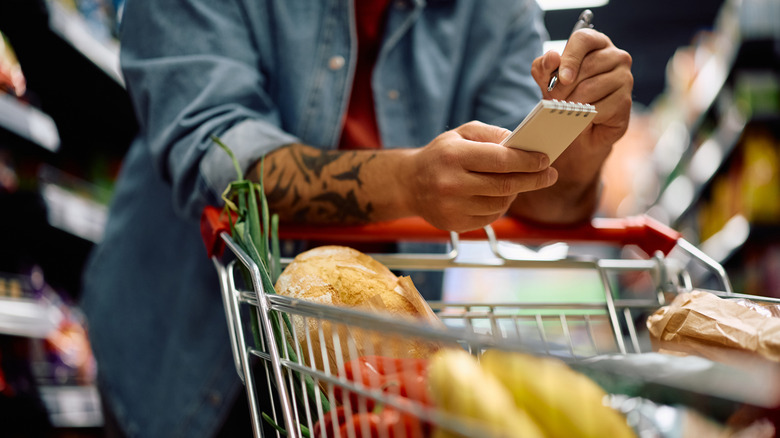 A tattooed person holding a notepad shopping list behind a full shopping cart in a grocery store