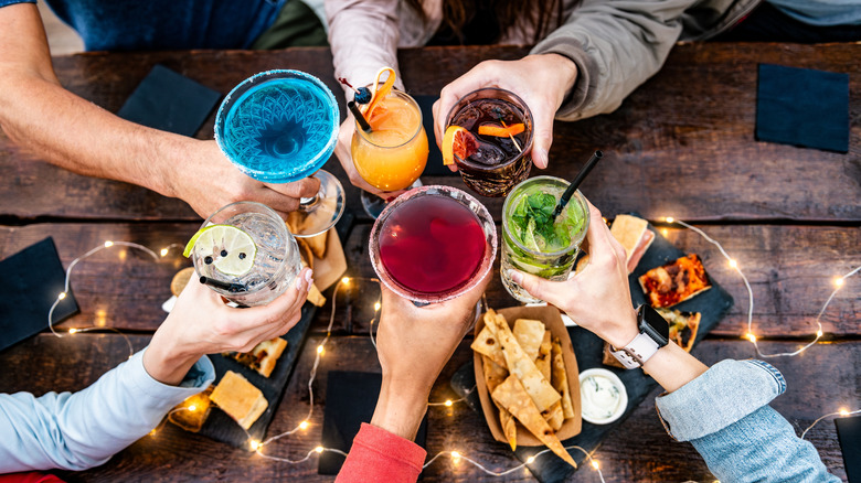 Six people's hands raise cocktail glasses in a toast over a wooden table with appetizers and lights