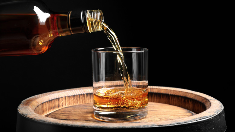 An unmarked bottle of whiskey being poured into a glass on top of a barrel