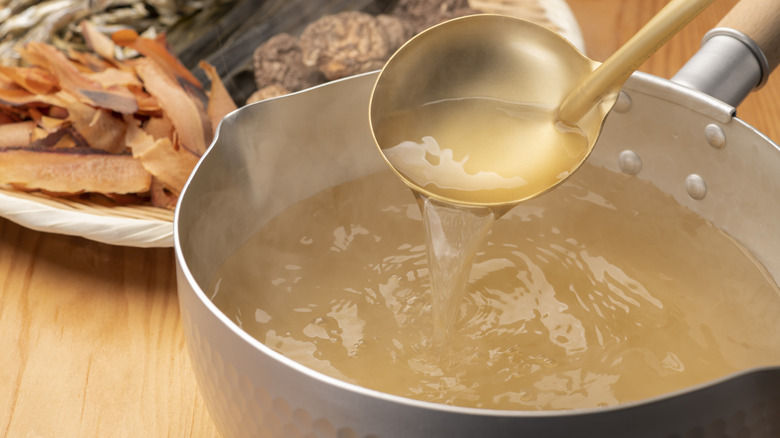 A ladle pouring a spoonful of seafood stock into a pot with herbs and dashi on a plate in the background