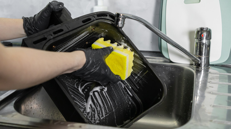 Gloved hands cleaning air fryer basket with soapy sponge in sink
