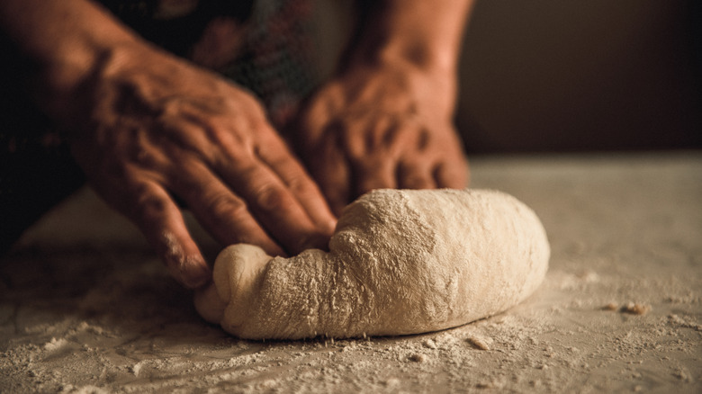 Two hands kneading a ball of bread dough on a surface with flour on it.