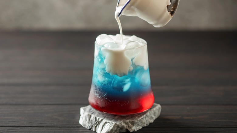 Person pouring cream into cherry blue dirty soda in glass