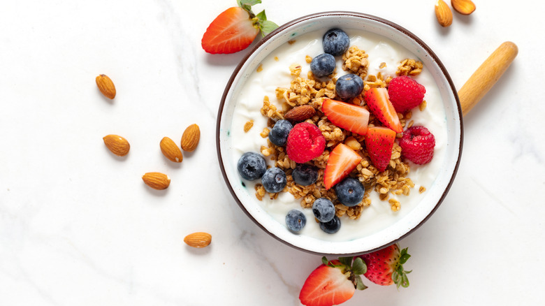 Bowl of yogurt with blueberries, strawberries, and granola near almonds on a white surface